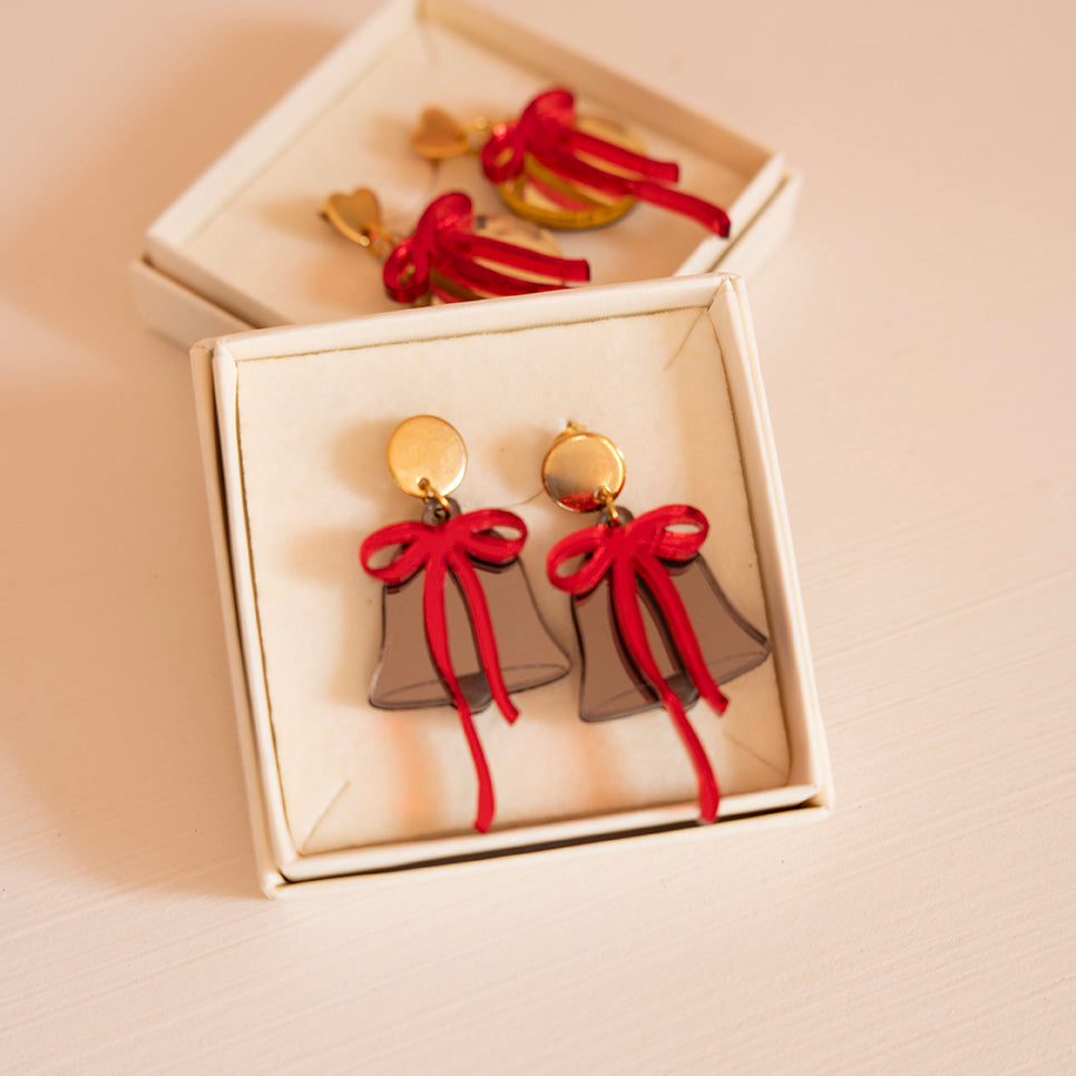 Dans une boîtes une paire de boucle d’oreilles représentant une cloche avec un noeud
attache ronde doré en métal
cloche en colori bronze et par dessus un noeud rouge asymétrique