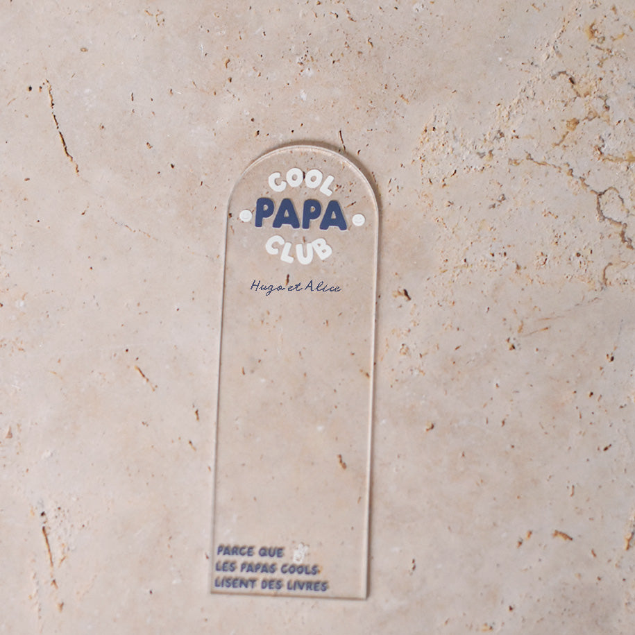 Marque-pages en plexi transparent découpe forme arche, texte blanc et bleu. Inscription dans l’arche sur 3 lignes « Cool Papa Club », 2 smiley entourent le mot « PAPA ». Les mots « cool » et « club » et les motifs sont inscrits en blanc, le reste du texte est en couleur.
En dessous est inscrit un prénoms (personnalisation du produit)
En bas du marque-page est inscrit « Parque les papas cools lisent des livres » disposé sur 3 lignes avec un dessin de main en « v »