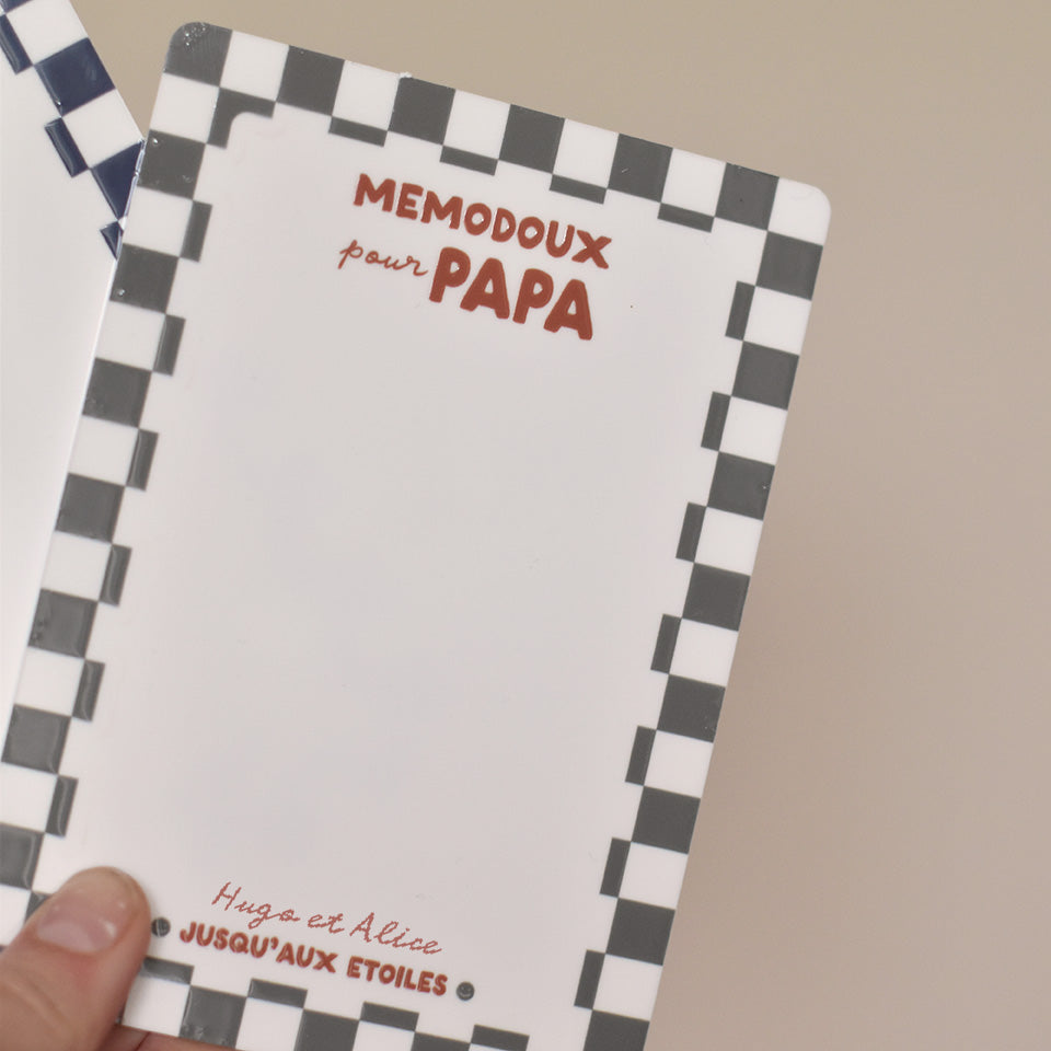 Ardoise effaçable en plexiglass blanc avec un cadre à motif damier vert, inscription centrée en haut « memodoux pour papa » couleur rouge terracotta, inscription centré en bas de deux prénoms « Hugo et Alice » et en dessous « jusqu’au étoiles » avec deux smileys sur les côtés