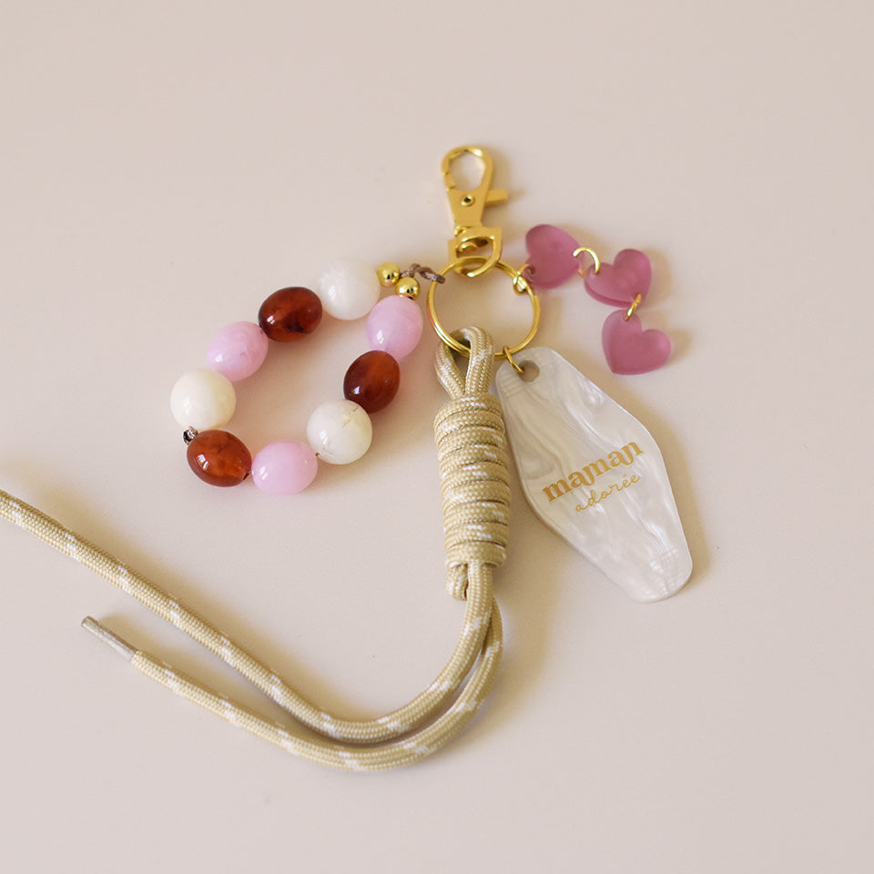 Grigris / bijoux de sac composé de 4 élèments sur un mousqueton doré élégant et un anneau porte-clé : une paracorde beige de 20 cm de long, trois petits cœurs roses attachés par des anneaux dorés, des perles acryliques aux tons blanc, rose et ambre, et un porte-clé style motel en plexiglass marbré blanc imprimé « Maman adorée » couleur doré