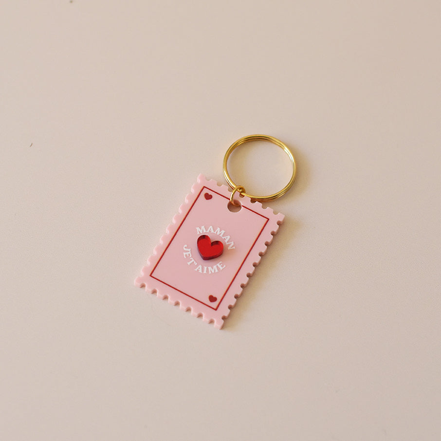 Porte-clés plexi coloré rose et rouge, design de carte à jour, forme de timbre et coeur au centre en relief, anneau doré. Inscription maman je t’aime