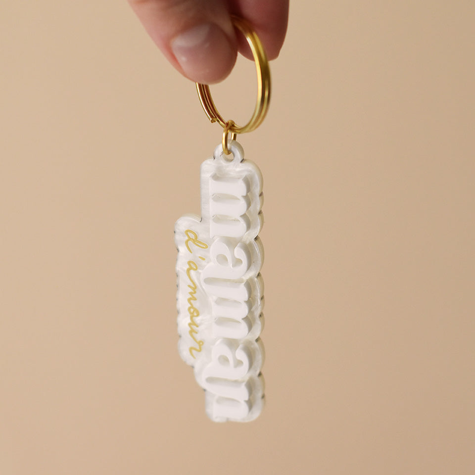 Porte-clés plexi coloré fond marbré blanc inscription « maman d’amour », « maman » en relief blanc et « d’amour » imprimé en doré, découpe à la forme du lettrage sur anneau doré