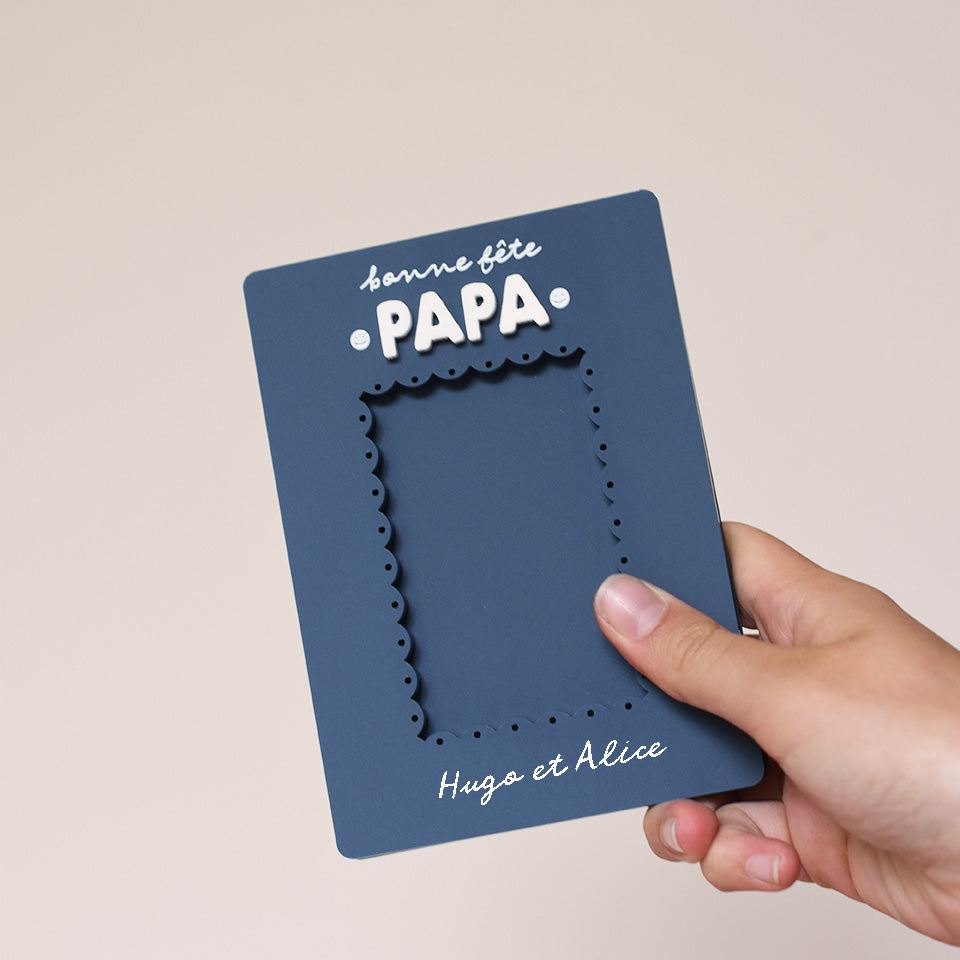 Cadre en plexi, format rectangulaire colors bleu « denim » et texte blanc. Découpe de l’ouverture au centre en feston 
Inscription en haut centré sur 2 lignes, ligne 1 « bonne fête », ligne 2 « Papa » en relief plexi blanc et sur les côtés du mot 2 smileys imprimés
Inscription en bas de deux prénoms