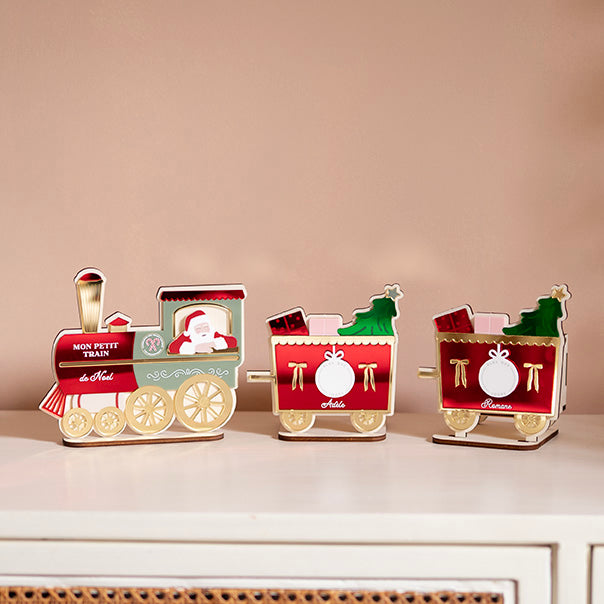 Décor en bois avec relief coloré 
Représentation d’un petit train 
Locomotive rouge, verte et doré avec père-noël accouché à la fenêtre
Derrière 2 wagons identiques : fond rouge avec détail doré, deux cadeaux et un sapin sorte de ceux-ci, Au centre un rond blanc, sur le coté 2 noeus dorés, et un prénom est inscrit