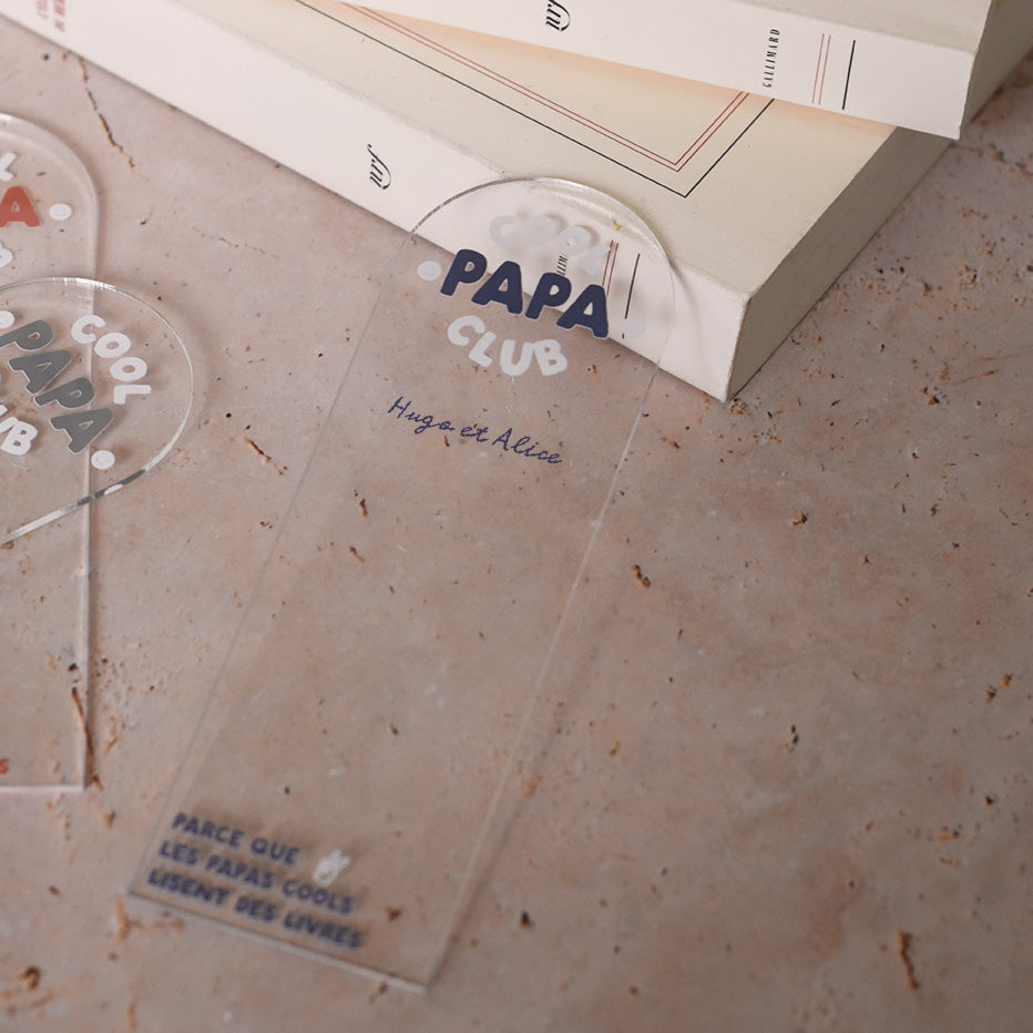 Marque-pages en plexi transparent découpe forme arche, texte blanc et bleu. Inscription dans l’arche sur 3 lignes « Cool Papa Club », 2 smiley entourent le mot « PAPA ». Les mots « cool » et « club » et les motifs sont inscrits en blanc, le reste du texte est en couleur.
En dessous est inscrit un prénoms (personnalisation du produit) 
En bas du marque-page est inscrit « Parque les papas cools lisent des livres » disposé sur 3 lignes avec un dessin de main en « v » 