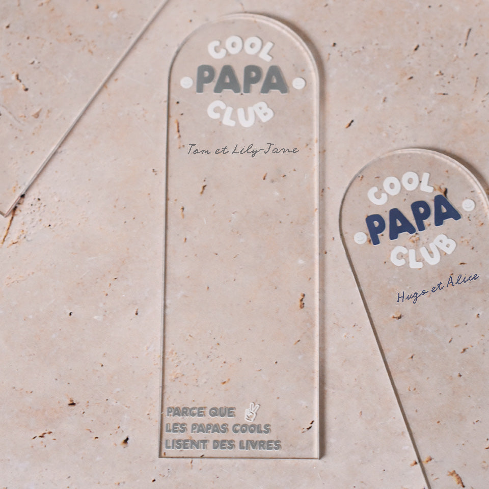 Marque-pages en plexi transparent découpe forme arche, texte blanc et coloré. Un modèle texte vert et blanc et un modèle texte bleu et blanc sont présentés.
Inscription dans l’arche sur 3 lignes « Cool Papa Club », 2 smiley entourent le mot « PAPA ».
En dessous est inscrit un prénoms (personnalisation du produit) 
En bas du marque-page est inscrit « Parque les papas cools lisent des livres » disposé sur 3 lignes avec un dessin de main en « v » 