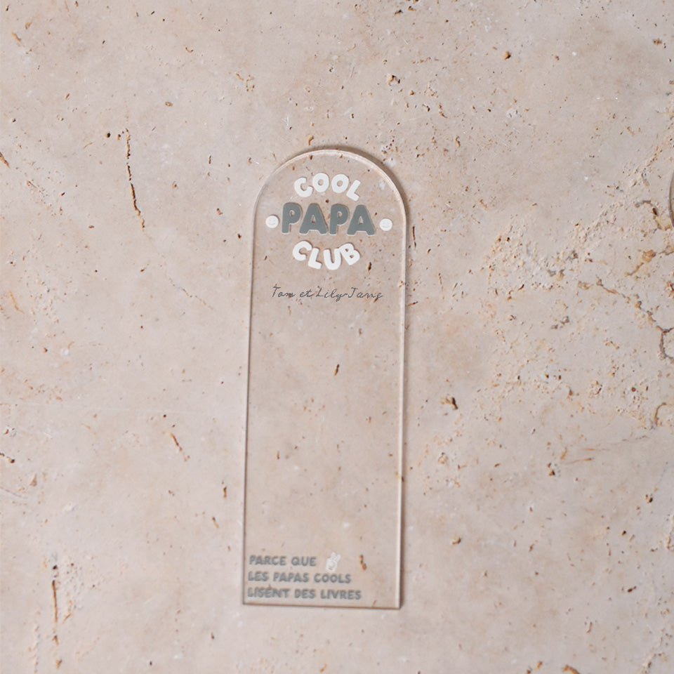 Marque-pages en plexi transparent découpe forme arche, texte blanc et vert. Inscription dans l’arche sur 3 lignes « Cool Papa Club », 2 smiley entourent le mot « PAPA ». Les mots « cool » et « club » et les motifs sont inscrits en blanc, le reste du texte est en couleur.
En dessous est inscrit un prénoms (personnalisation du produit) 
En bas du marque-page est inscrit « Parque les papas cools lisent des livres » disposé sur 3 lignes avec un dessin de main en « v » 