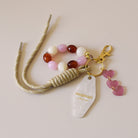 Grigris / bijoux de sac composé de 4 élèments sur un mousqueton doré élégant et un anneau porte-clé : une paracorde beige de 20 cm de long, trois petits cœurs roses attachés par des anneaux dorés, des perles acryliques aux tons blanc, rose et ambre, et un porte-clé style motel en plexiglass marbré blanc imprimé « Maman adorée » couleur doré