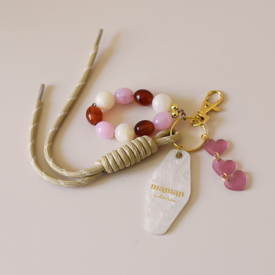Grigris / bijoux de sac composé de 4 élèments sur un mousqueton doré élégant et un anneau porte-clé : une paracorde beige de 20 cm de long, trois petits cœurs roses attachés par des anneaux dorés, des perles acryliques aux tons blanc, rose et ambre, et un porte-clé style motel en plexiglass marbré blanc imprimé « Maman adorée » couleur doré