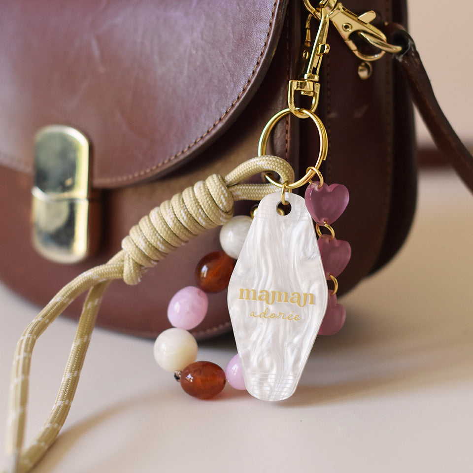 Grigris / bijoux de sac composé de 4 élèments sur un mousqueton doré élégant et un anneau porte-clé : une paracorde beige de 20 cm de long, trois petits cœurs roses attachés par des anneaux dorés, des perles acryliques aux tons blanc, rose et ambre, et un porte-clé style motel en plexiglass marbré blanc imprimé « Maman adorée » couleur doré
Photo du produit sur sac