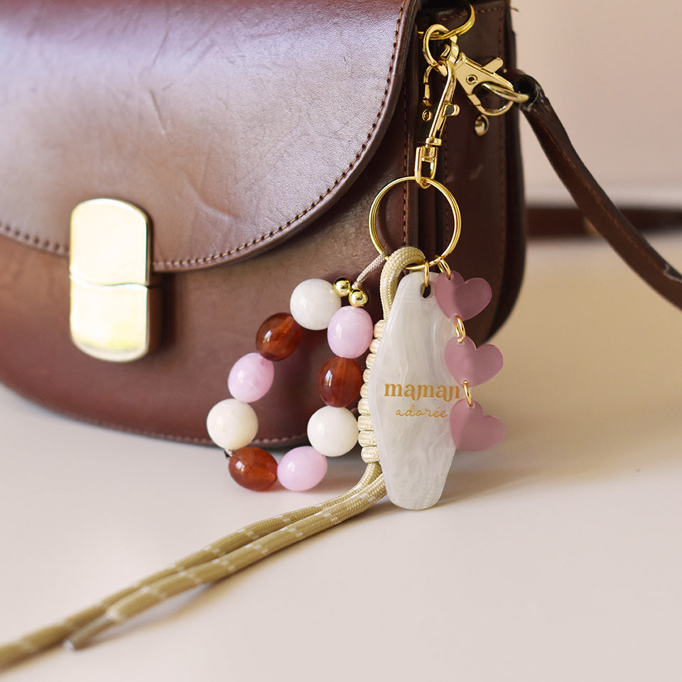 Grigris / bijoux de sac composé de 4 élèments sur un mousqueton doré élégant et un anneau porte-clé : une paracorde beige de 20 cm de long, trois petits cœurs roses attachés par des anneaux dorés, des perles acryliques aux tons blanc, rose et ambre, et un porte-clé style motel en plexiglass marbré blanc imprimé « Maman adorée » couleur doré
Photo du produit sur sac
