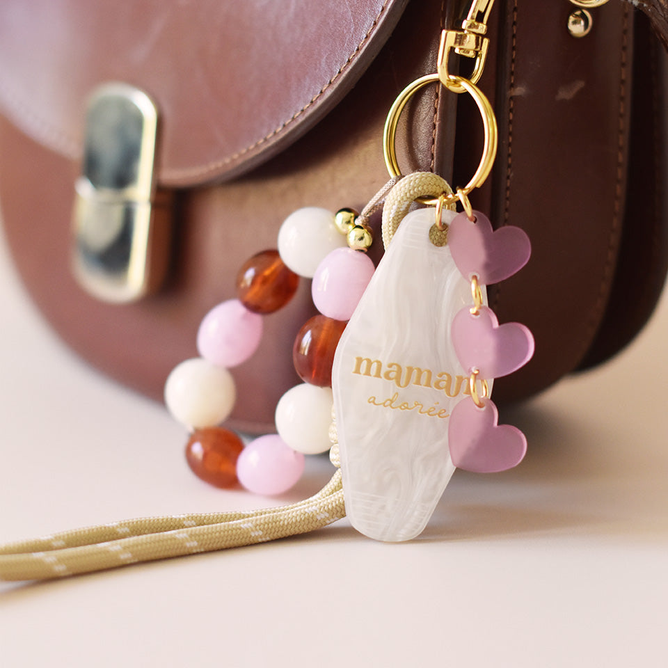 Grigris / bijoux de sac composé de 4 élèments sur un mousqueton doré élégant et un anneau porte-clé : une paracorde beige de 20 cm de long, trois petits cœurs roses attachés par des anneaux dorés, des perles acryliques aux tons blanc, rose et ambre, et un porte-clé style motel en plexiglass marbré blanc imprimé « Maman adorée » couleur doré
Photo du produit sur sac - Focus porte-clés