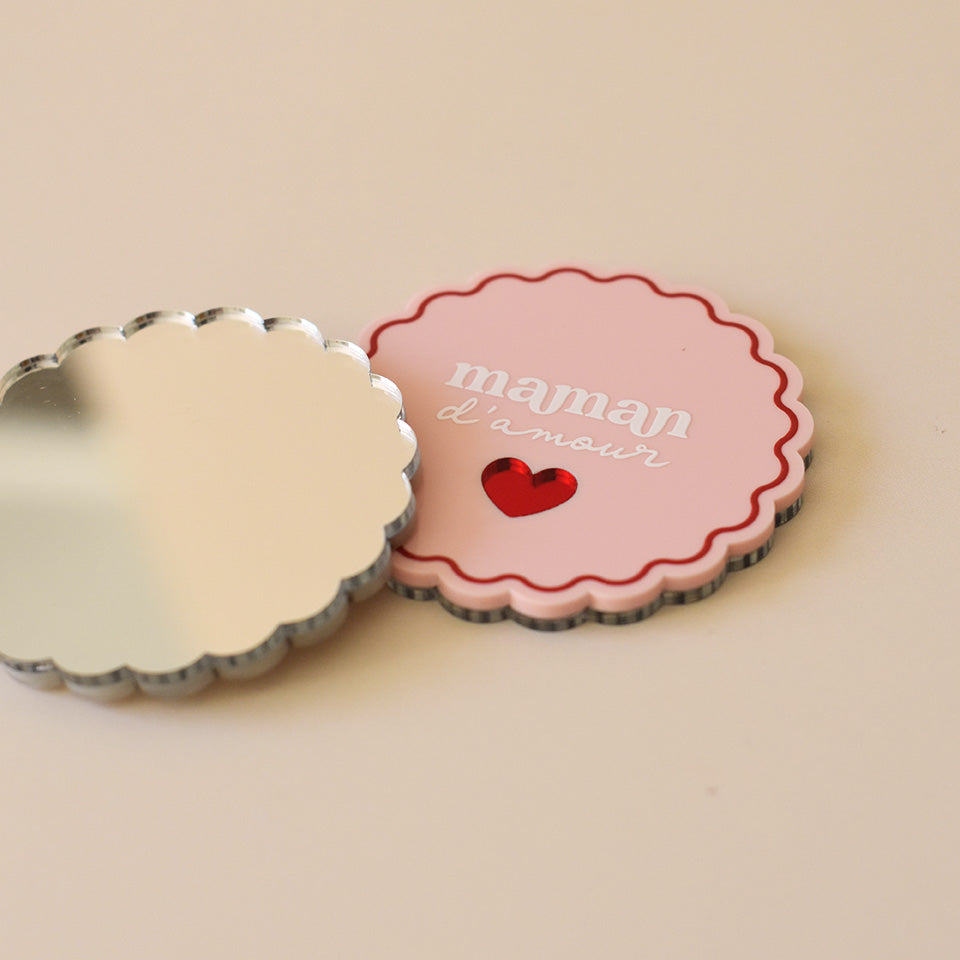 Miroir de poche forme rond festonné avec impression contour, inscription « maman d’amour » et coeur en plexi miroir incrusté au centre. Coloris face rose et rouge (fond plexi rose, contour rouge, texte blanc, coeur en plexi miroir rouge) / verso miroir argent