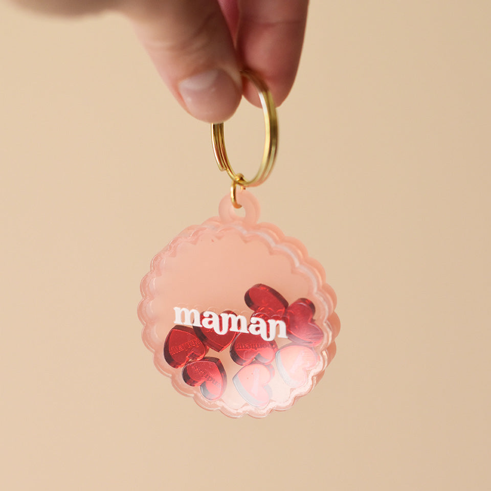 Porte-clés en plexiglass transparent et coloré rose sur anneaux dorés. Il a une forme de cercle festonné et contient 8 mini découpes en forme de cœur rouge qui bougent librement à l’intérieur. Ces découpes sont gravées de mots associés à la maternités «  tendresse, amour, bisous.. »