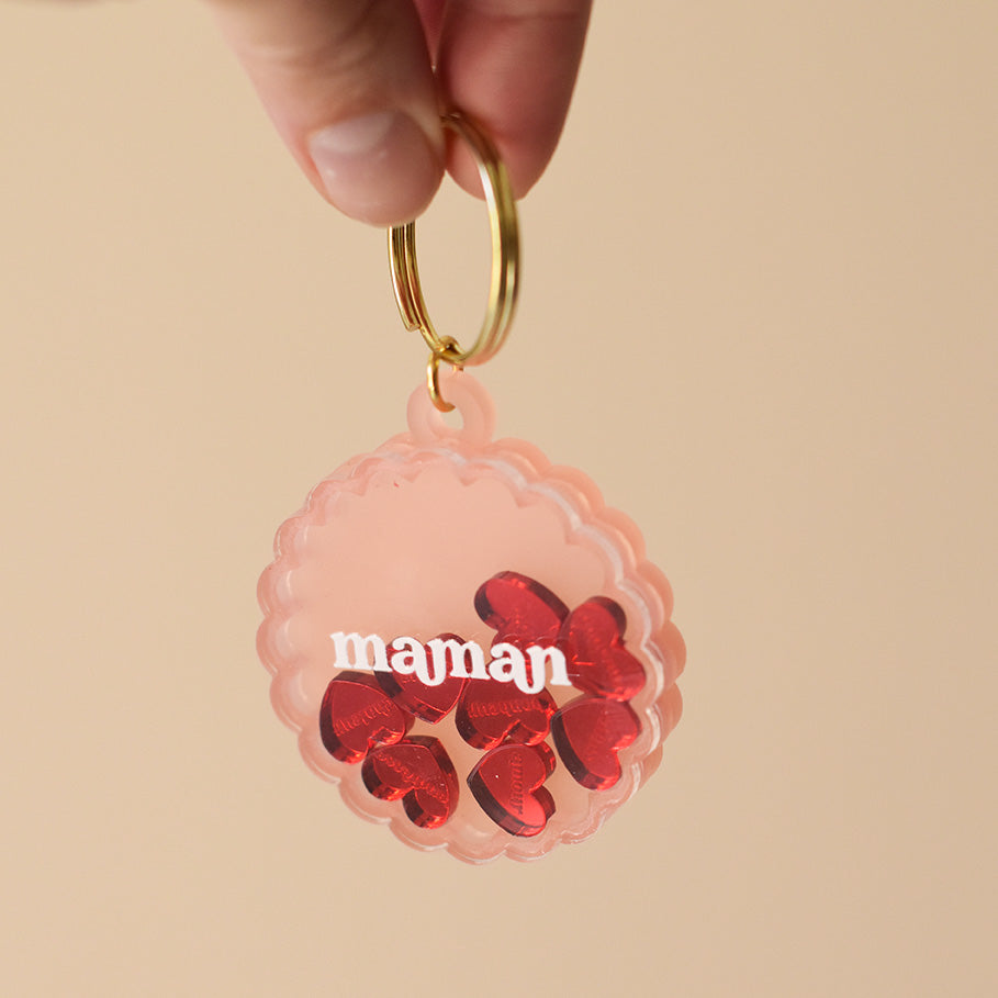 Porte-clés type en plexiglass transparent et coloré rose sur anneaux dorés. Il a une forme de cercle festonné et contient 8 mini découpes en forme de cœur rouge qui bougent librement à l’intérieur. Ces découpes sont gravées de mots associés à la maternités «  tendresse, amour, bisous.. »