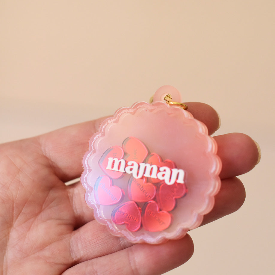 Porte-clés type en plexiglass transparent et coloré rose sur anneaux dorés. Il a une forme de cercle festonné et contient 8 mini découpes en forme de cœur rouge qui bougent librement à l’intérieur. Ces découpes sont gravées de mots associés à la maternités «  tendresse, amour, bisous.. »
Photo plan rapproché