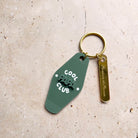 Découpes plexiglass assemblées sur anneau et anneau brisé dorés 
Se compose de deux élément.

Elément 1 :  découpe forme motel en plexi mat couleur vert « pin » et texte blanc
inscription « cool papa club » sur 3 ligne, « papa » en relief ton sur ton, « cool » et « club » imprimé en blanc
+ 2 smileys entourent l’inscription « papa »
Elément 2 : une découpe en plexi miroir or forme rectangle arrondi et gravé avec une  inscription de prénoms 