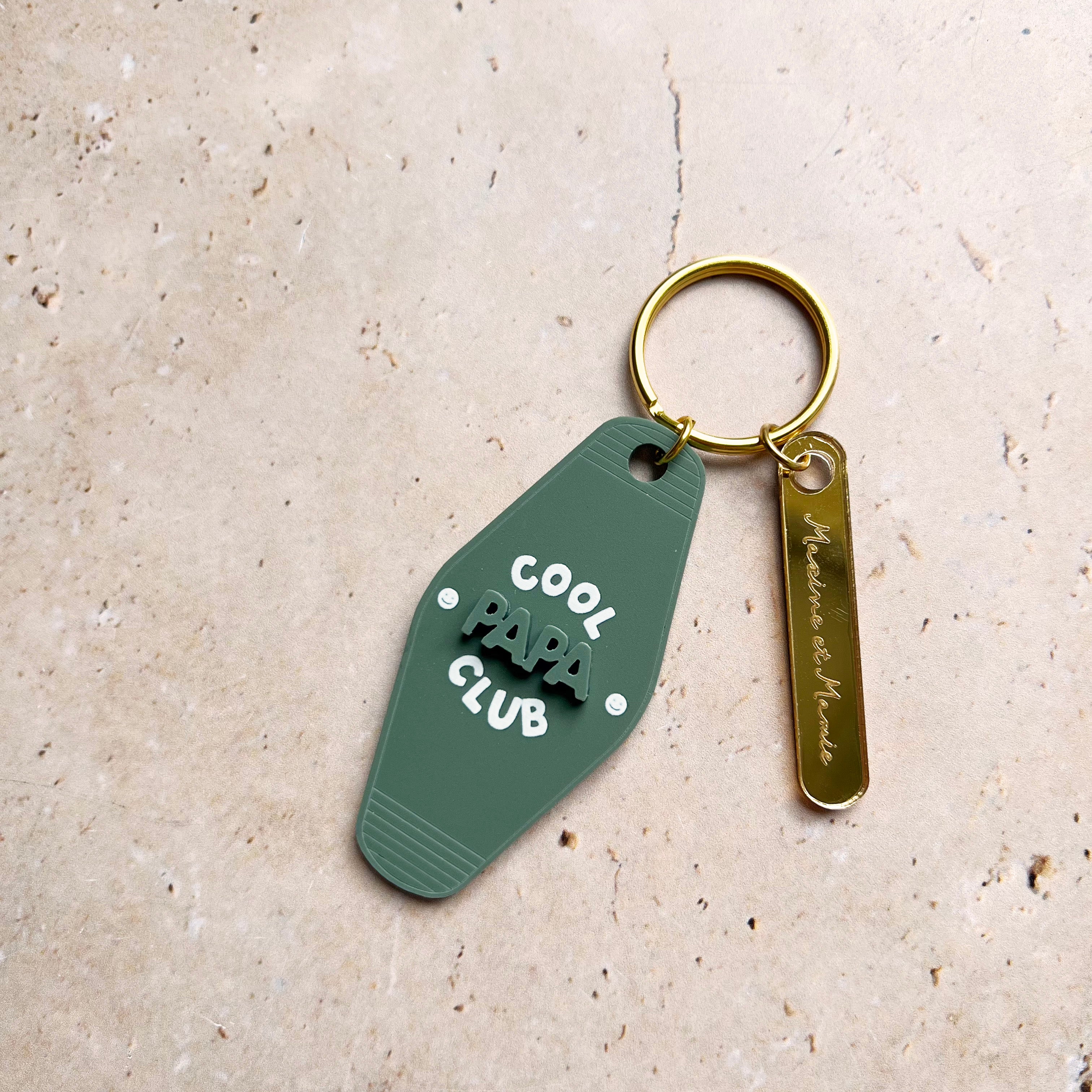 Découpes plexiglass assemblées sur anneau et anneau brisé dorés 
Se compose de deux élément.

Elément 1 :  découpe forme motel en plexi mat couleur vert « pin » et texte blanc
inscription « cool papa club » sur 3 ligne, « papa » en relief ton sur ton, « cool » et « club » imprimé en blanc
+ 2 smileys entourent l’inscription « papa »
Elément 2 : une découpe en plexi miroir or forme rectangle arrondi et gravé avec une  inscription de prénoms 