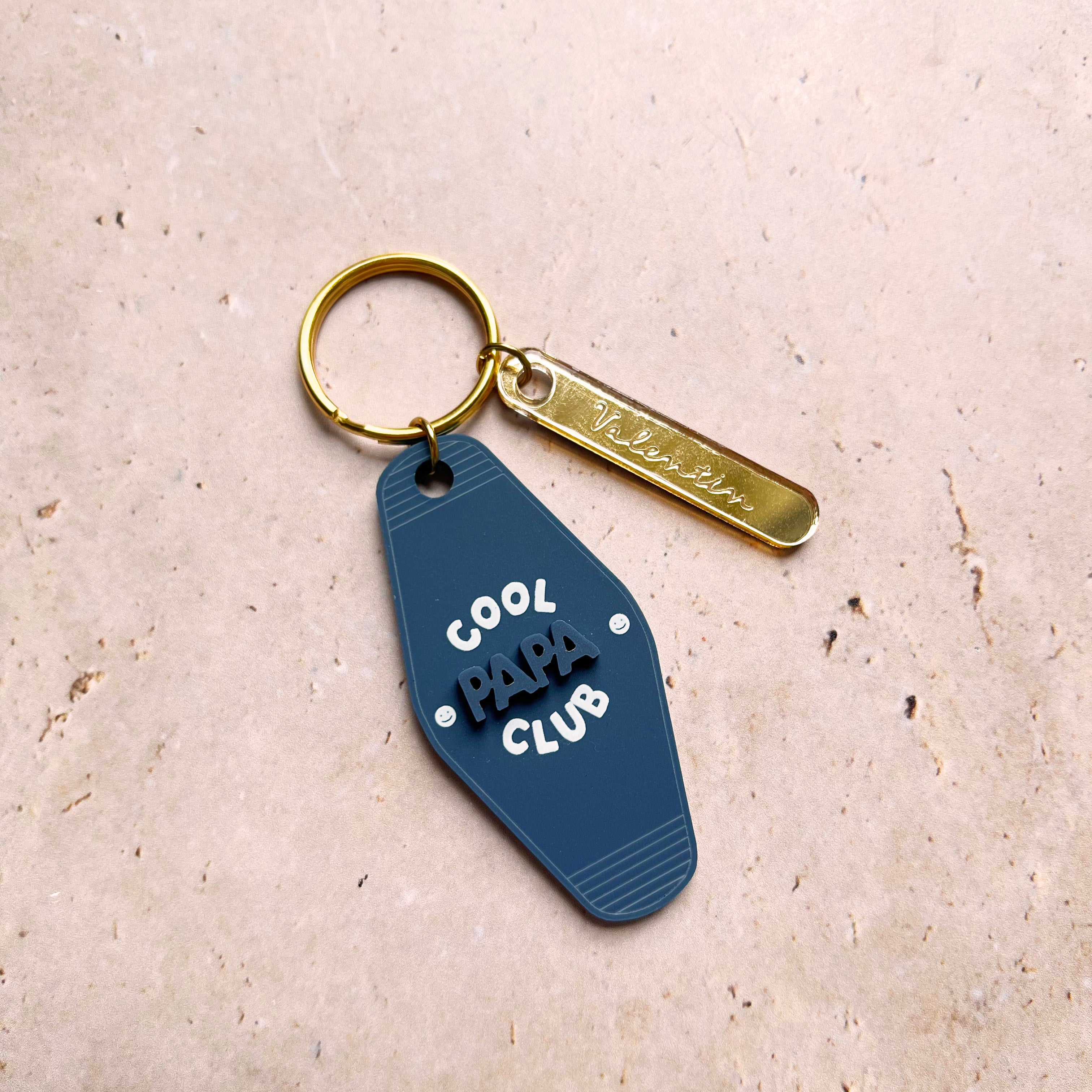 Découpes plexiglass assemblées sur anneau et anneau brisé dorés 
Se compose de deux élément.

Elément 1 :  découpe forme motel en plexi mat couleur bleu « denim » et texte blanc
inscription « cool papa club » sur 3 ligne, « papa » en relief ton sur ton, « cool » et « club » imprimé en blanc
+ 2 smileys entourent l’inscription « papa »
Elément 2 : une découpe en plexi miroir or forme rectangle arrondi et gravé avec une  inscription de prénoms 